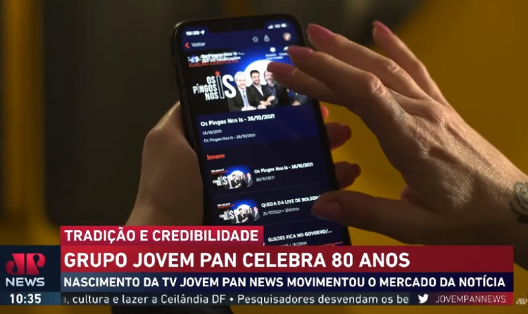Frame de reportagem sobre os 80 anos da Jovem Pan mostra mão segurando um celular em que vídeo do Pingos nos Is é exibido