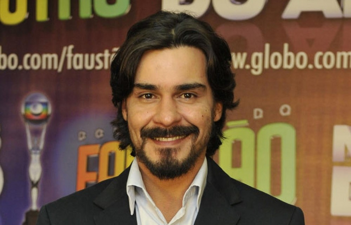 Ator André Gonçalves sorri em frente a painel da Globo