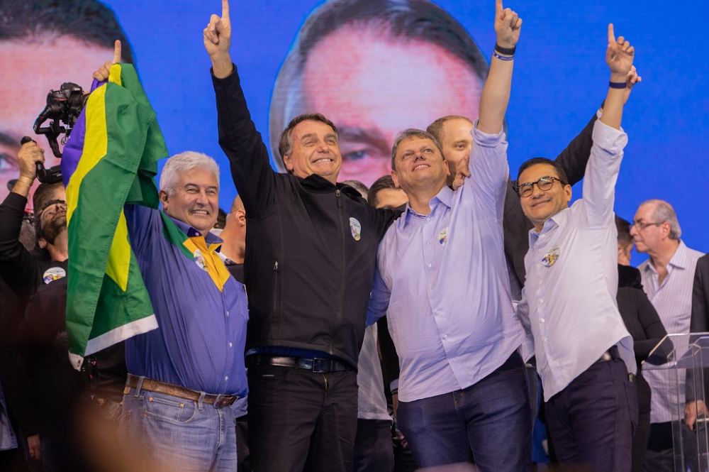 Marcos Pontes, segurando uma bandeira, Bolsonaro e Tarcísio, os três em pé, abraçados e apontando para o alto
