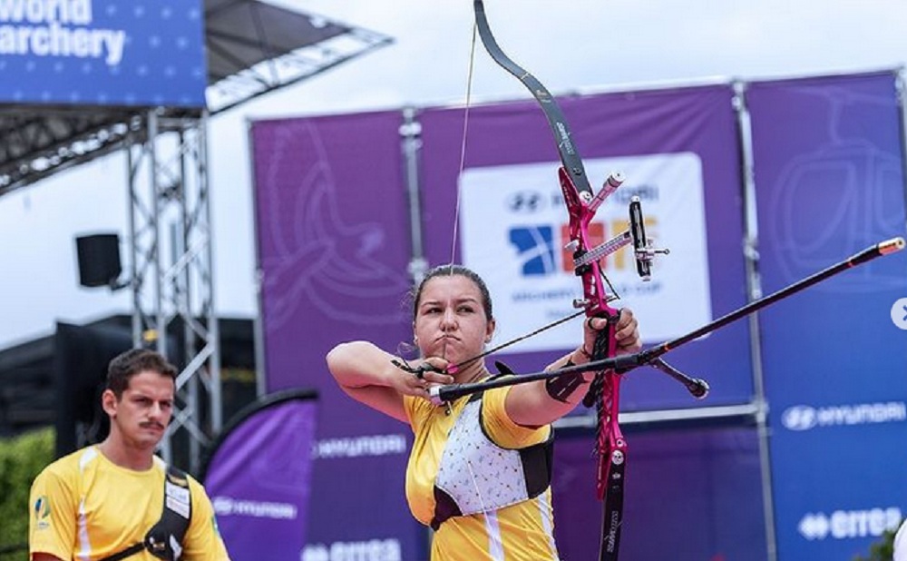 Atleta brasileira atira com arco; companheiro de dupla observa ao fundo