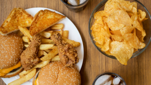 Mesa cheia de comidas gordurosas como X-burger, batata frita, frango frito e batata chips