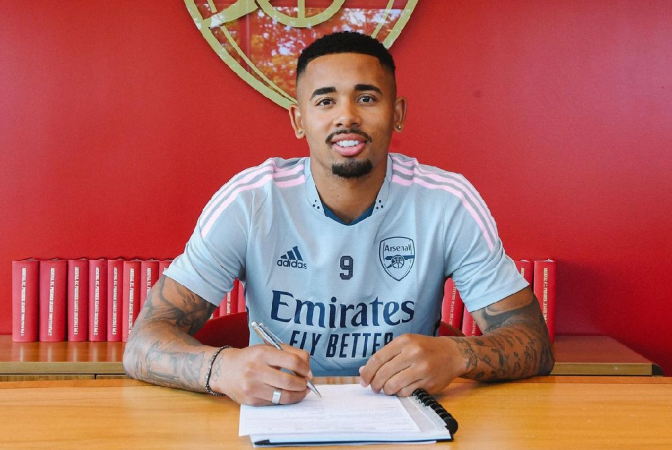 Gabriel Jesus sentado à mesa assina contrato com a camisa do Arsenal , com parede vermelha com sóimbolo do Arsenal atrás