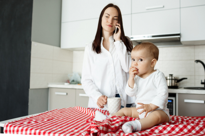 Mãe tomando café e falando ao telefone enquanto o bebê come na mesa da cozinha