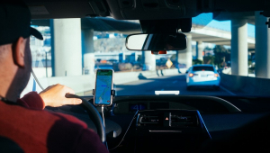 Imagem mostra um motorista de boné, de costas para a câmera; há foco no celular que está com GPS ligado