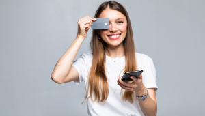 Jovem mulher branca sorri com o celular na mão esquerda e o cartão na mão direita, tampando o olho direito