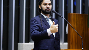O deputado David Miranda, de terno, gravata e cabelo com tranças, faz sinal de joia em plenário