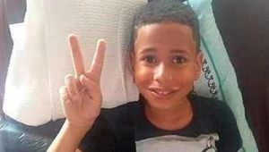 Menino de 10 anos deitado na cama faz sinal de V com a mão direita