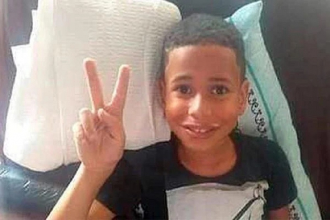 Menino de 10 anos deitado na cama faz sinal de V com a mão direita