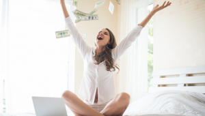 Mulher com computador na cama joga dólares para cima e abre os braços, mostrando felicidade