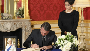 O presidente Jair Bolsonaro, sentado, assina o livro de condolências da rainha Elizabeth II; sua mulher Michelle está ao seu lado, toda de preto