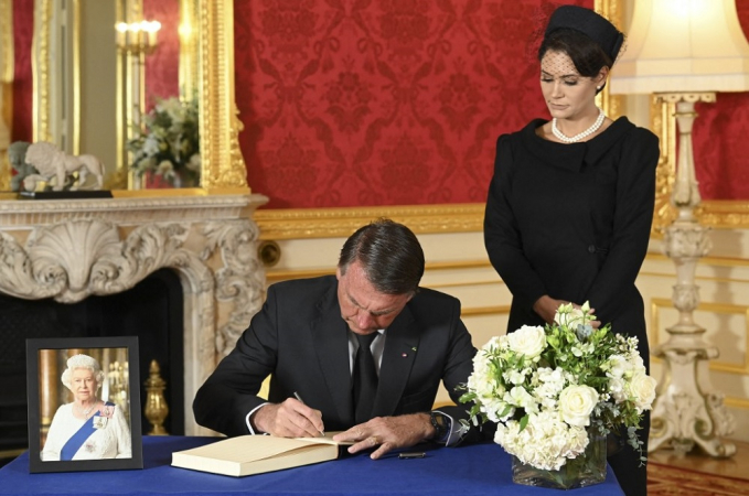 O presidente Jair Bolsonaro, sentado, assina o livro de condolências da rainha Elizabeth II; sua mulher Michelle está ao seu lado, toda de preto