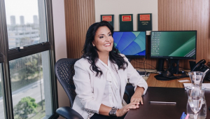 Mulher branca e de cabelo preto, na faixa dos 40/50 anos, sorri sentada à mesa de seu escritório