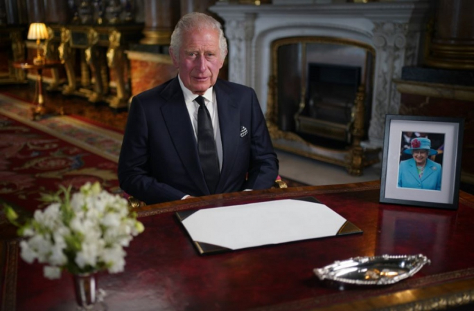 King Charles III seated at a table decorated with a photo of his mother