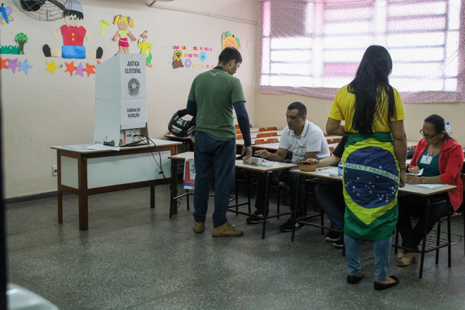 Um homem de verde e uma mulher com a bandeira do Brasil enrolada na cintura falam com mesários antes de votar