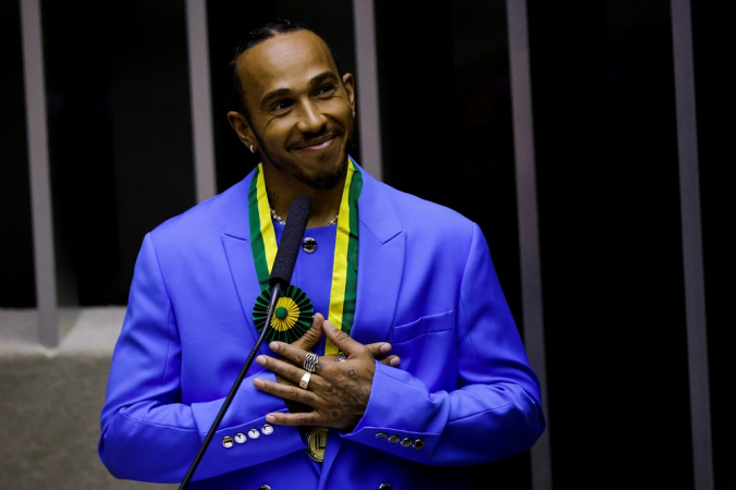 Vestido de terno azul, Lewis Hamilton reage após receber o título de cidadão honorário do Brasil na Câmara dos Deputados