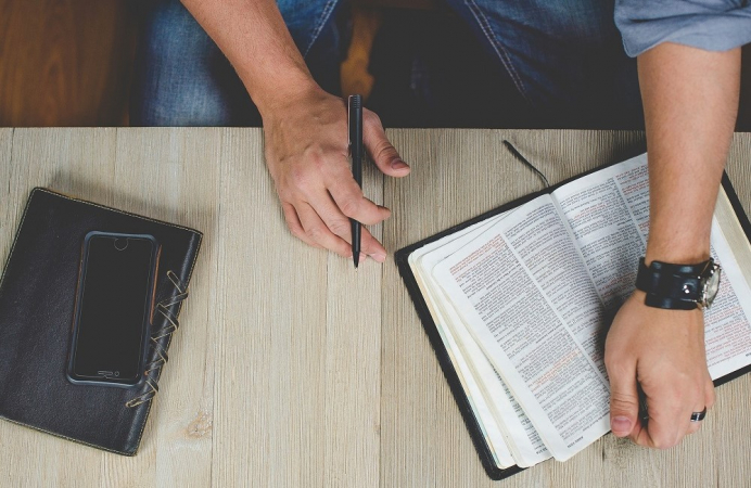 Mãso sehurando caneta (esquerda) e Bíblia (direita) em mesa de madeira com celular e agenda