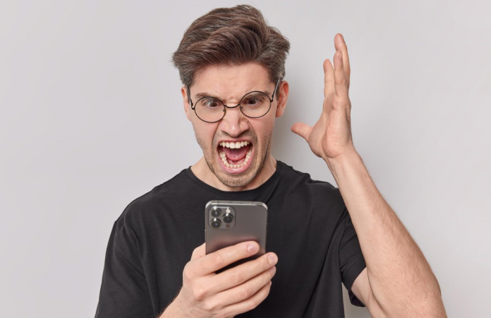Homem irritado grita com raiva e mantém a palma levantada olhando para smartphone