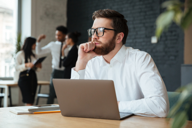 Homem branco de negócios, com barba e óculos, concenrado em frente a um notebook emnquanto três amigos conversam no fundo