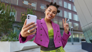 Garota atraente com roupa elegante cria conteúdo de influência e fa sinal da paz em pose de câmera frontal do smatphone e sorri em local urbano