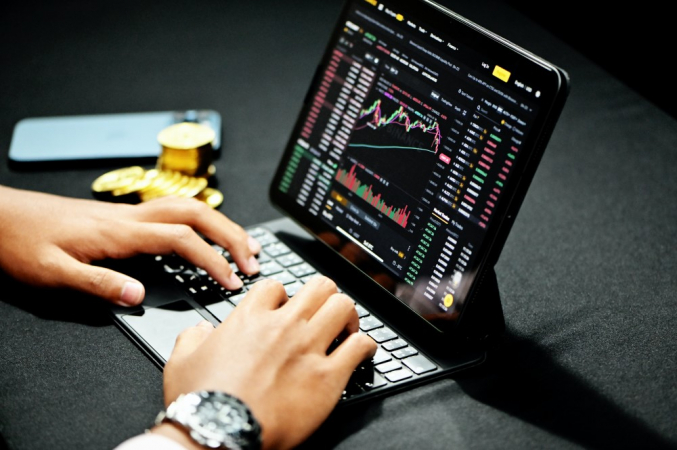 Homem digita em notebook com gráficos de investimentos, em superfície cinza, e exibe relógio no braço direito (só os braços aparecem); ao lado esquerdo, aparecem moedas e um celular