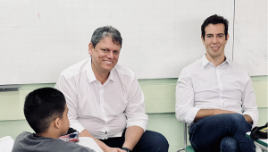 Tarcísio Gomes de Freitas e Renato Feder sentados em sala de aula, de costas para lousa, de frente pata uma criança, que na imagem aparece de costas