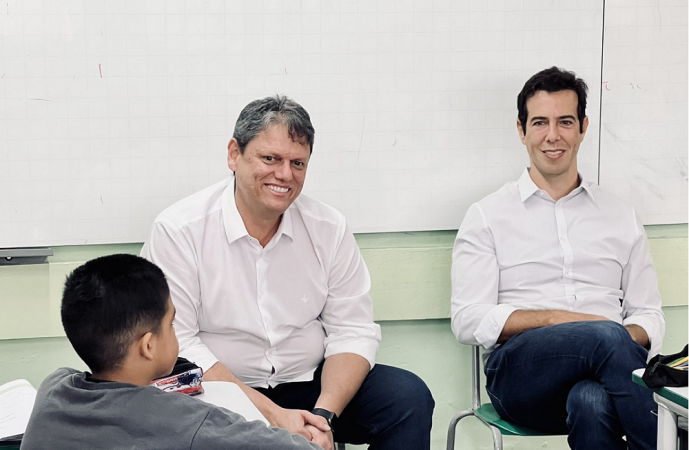 Tarcísio Gomes de Freitas e Renato Feder sentados em sala de aula, de costas para lousa, de frente pata uma criança, que na imagem aparece de costas