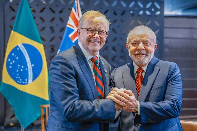 Com as bandeiras de Brasil e Austrália atrás, Anthony Albanese e Lula apertam as mãos e sorriem