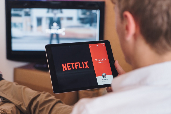 Homem na sala, sentado no sofá, segurando tablet na Netflix enquanto TV está ligada