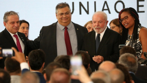 Dino e Lula muito próximos e cercados de vários pessoas em fgrente a uma parece onde está escrito União