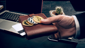 Homem segura carteira com bitcoins no topo, perto de mesa com computador
