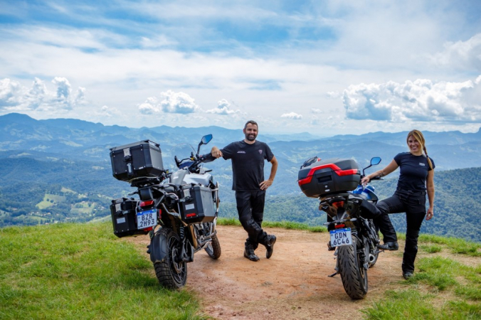 Os apresentadores Karina Simões e Edu Bernasconi ao lado de suas motos em uma montanha, de onde se vê o céu azul e uma paisagem com morros e árvores