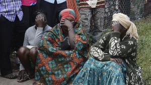 Pessoas sentadas no meio fio com a mão no rosto lamentam ataque em Uganda