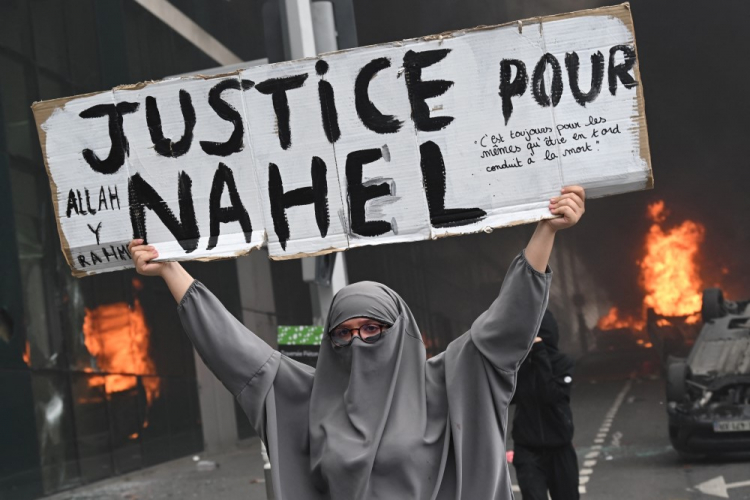 Um participante segura uma faixa com os dizeres "Justiça para Nahel" enquanto carros queimam na rua no final de uma marcha em homenagem a um motorista adolescente morto a tiros