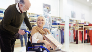 Idoso carrega idosa em cadeira de rodas em centro de compras
