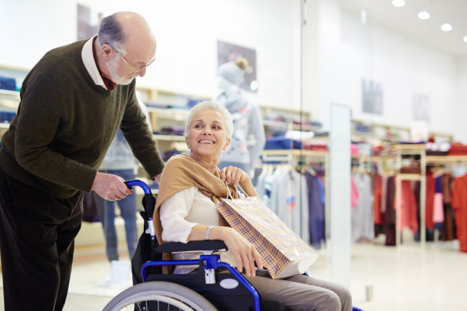 Idoso carrega idosa em cadeira de rodas em centro de compras
