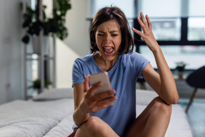 Mulher frustrada gritando com o telefone enquanto está sentada na cama e lendo a mensagem de texto que recebeu