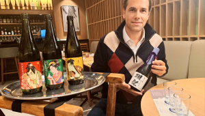 Empresário sentado em mesa de seu restaurante com saquês segura uma garrafa da bebida