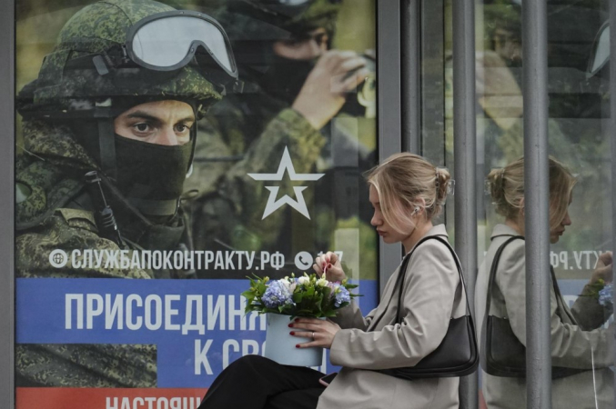 Uma mulher russa está sentada em um ponto de ônibus em frente a um cartaz anunciando o recrutamento militar mostrando soldados russos e a inscrição 'Junte-se ao seu. Trabalho de verdade.