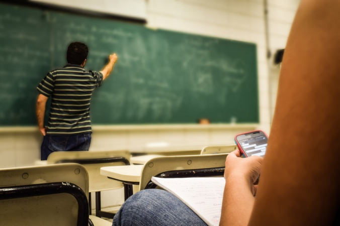 Estudante usa celular enquanto professor escreve na lousa
