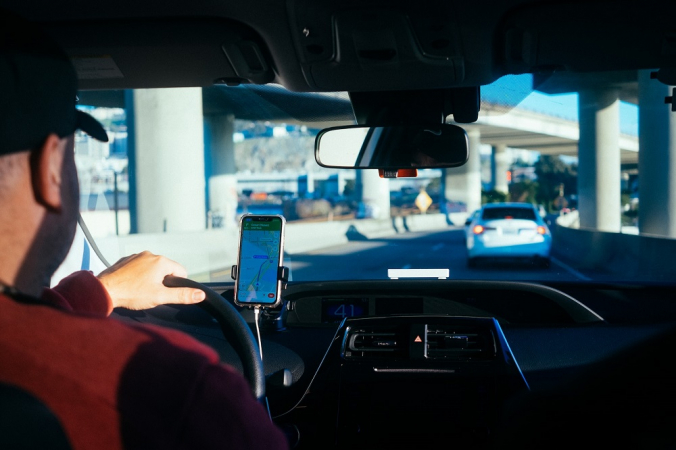 Visto de trás, Homem de boné dirige carro com auxílio de app de trajetos no celular