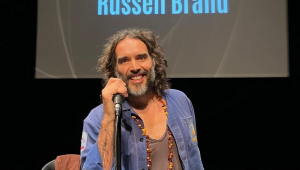 O ator Russel Brand num palco com seu nome em painel atrás