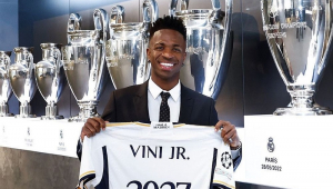 Sorrindo, Vinicius Júnior exibe a camisa do Real com o número 2027 em frente aos troféus da Champions que o clube conquistou