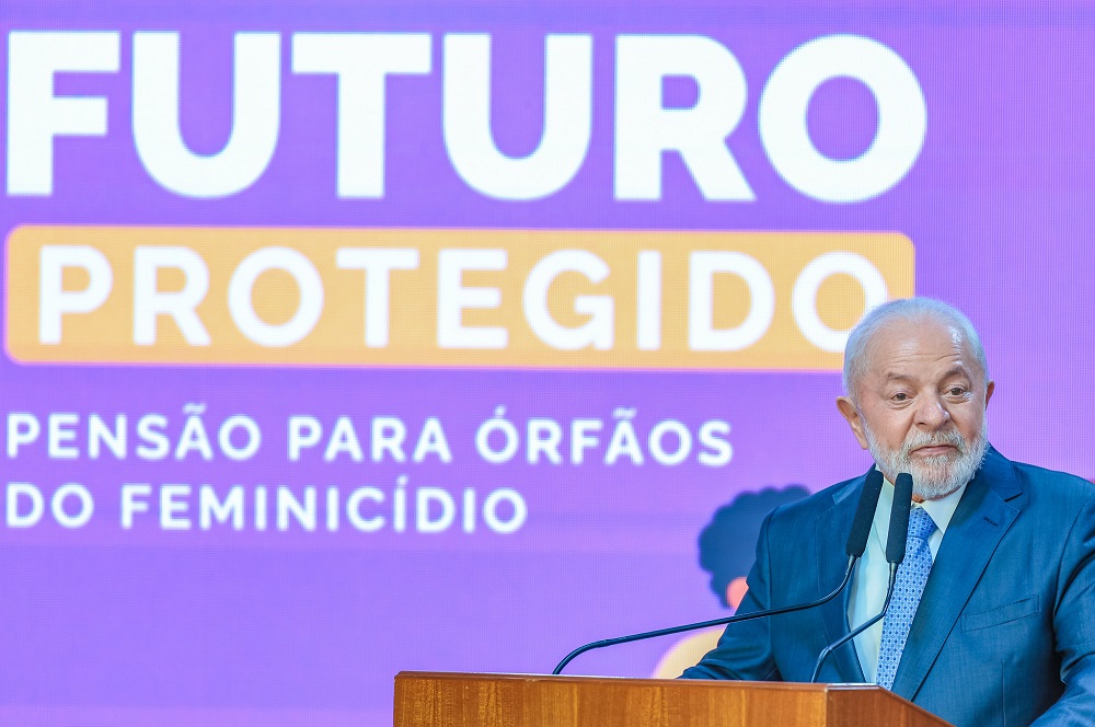 Lula em frente a uma parede azul onde se lê "Futuro protegido: pensão para órfãos de feminicídio"