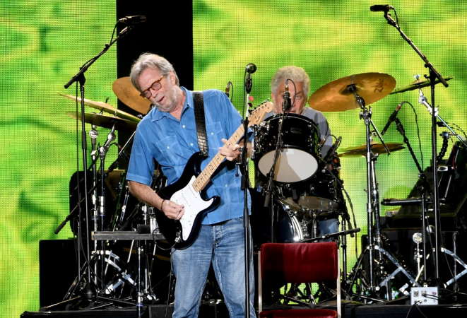 Eric Clapton toca guitarra com bateria e fundo verde no plano de fundo