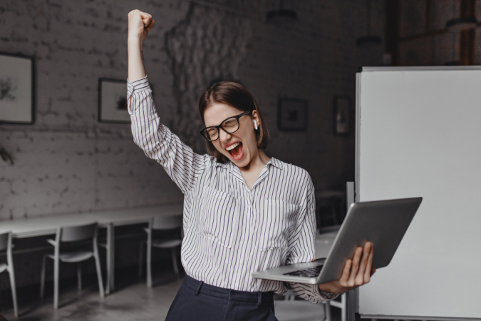 Mulher de negócios de óculos, blusa listrada e com o laptop na mão está feliz com o sucesso