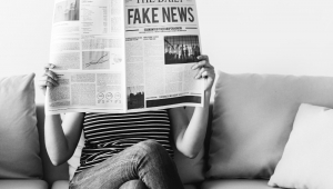Mulher abre jornal , qjue lhe cobre a cara, com o título fake news na manchete