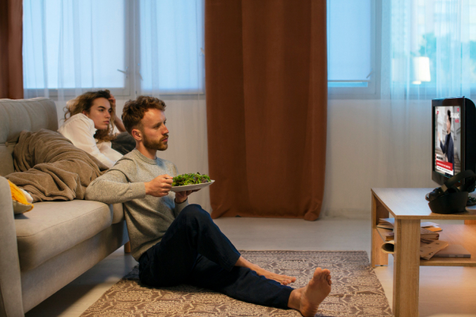 Mulher no sofá, homem comendo no chão, ambos vendo TV