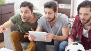 Três homens, vendo jogo na TV, sendo que um segura um tablet e outro uma bola