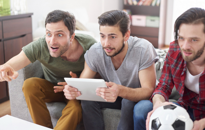 Três homens, vendo jogo na TV, sendo que um segura um tablet e outro uma bola