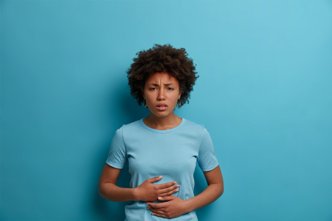 Mulher afro descontente sente desconforto no estômago, mantém as palmas das mãos na barriga, tem cólicas menstruais, veste uma camiseta casual, comeu comida estragada, isolada na parede azul, expressa emoções negativas com imitação